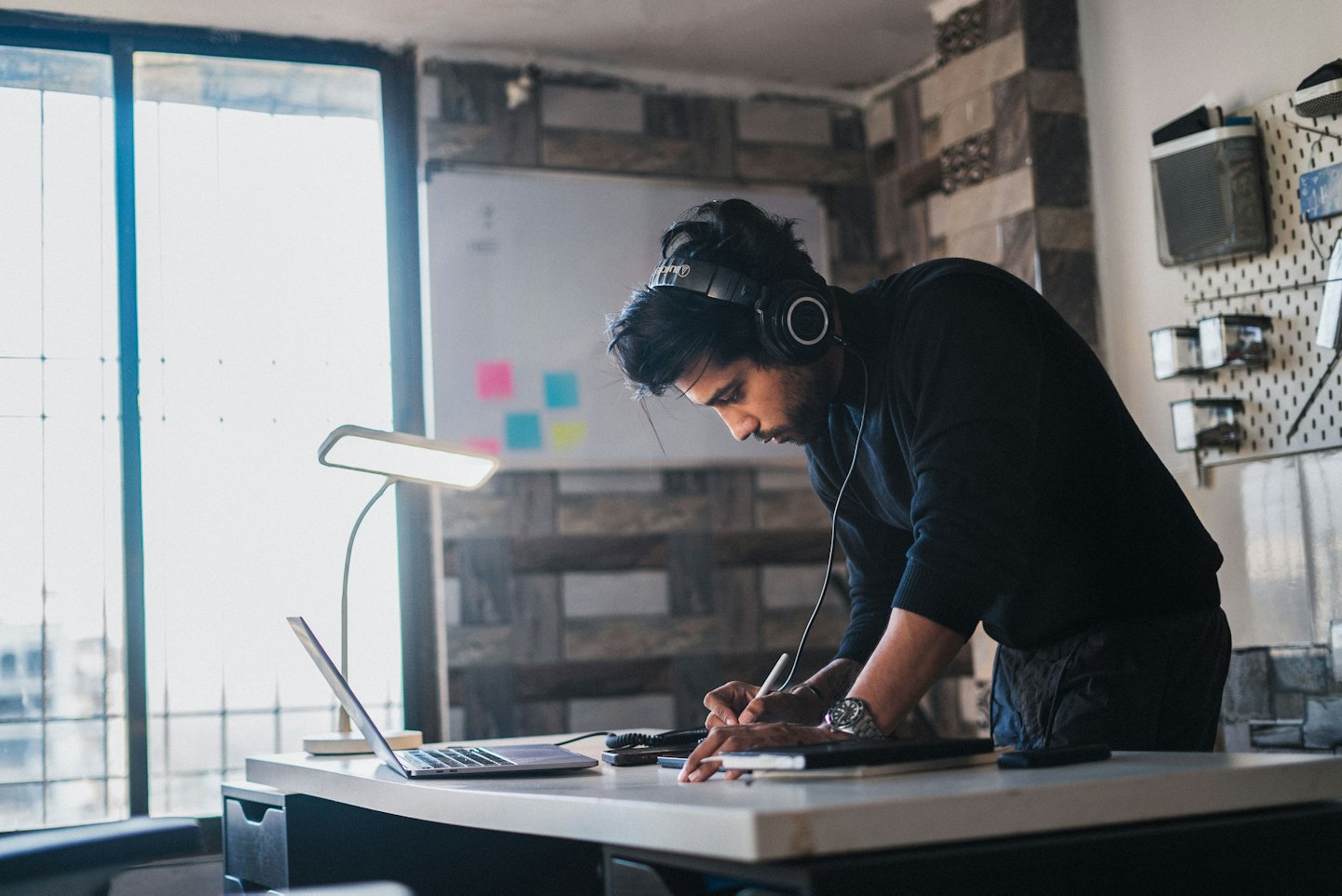 Man with headphones draws on tablet at desk with laptop.